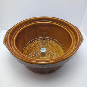 May include: A round, brown ceramic slow cooker insert with a glossy finish and vertical ribbed detailing on the exterior. Inside, a circular silver-toned metal wire rack is positioned at the base, featuring a small center plate embossed with the word RIVAL.