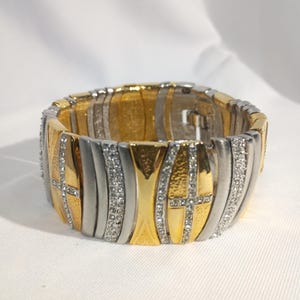 Puede incluir: Un brazalete ancho en tonos dorados y plateados con un diseño geométrico. El brazalete presenta segmentos dorados y plateados alternos, algunos adornados con pequeños cristales brillantes. Las secciones doradas tienen un acabado texturizado, agregando interés visual a la pieza.