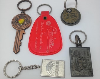 May include: Five assorted keychains including a copper-toned key with a crest, a red teardrop-shaped Chrysler Plymouth tag, a circular AutoZone gauge, a rectangular Ford keychain with a car illustration, and a silver square keychain with text.