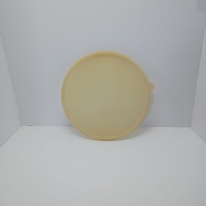 May include: A circular, light beige plastic lid with a smooth, flat surface and a small, protruding tab on the right side for easy removal. The lid features a slightly raised outer rim and a uniform, matte finish across its entire circular structure.