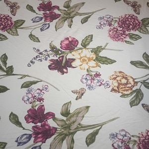 Könnte beinhalten: Ein weißer Stoff mit einem sich wiederholenden Muster aus verschiedenen Blumen in den Farben Dunkelmagenta, Gelb und Violett, ergänzt durch grüne Blätter und kleine braune Schmetterlinge, die über die Textiloberfläche verteilt sind.