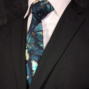 May include: A dark navy tie with a pattern of teal and yellow butterflies set against green leaves. The tie is knotted and worn with a white shirt and a black suit jacket. The image has a formal appearance.