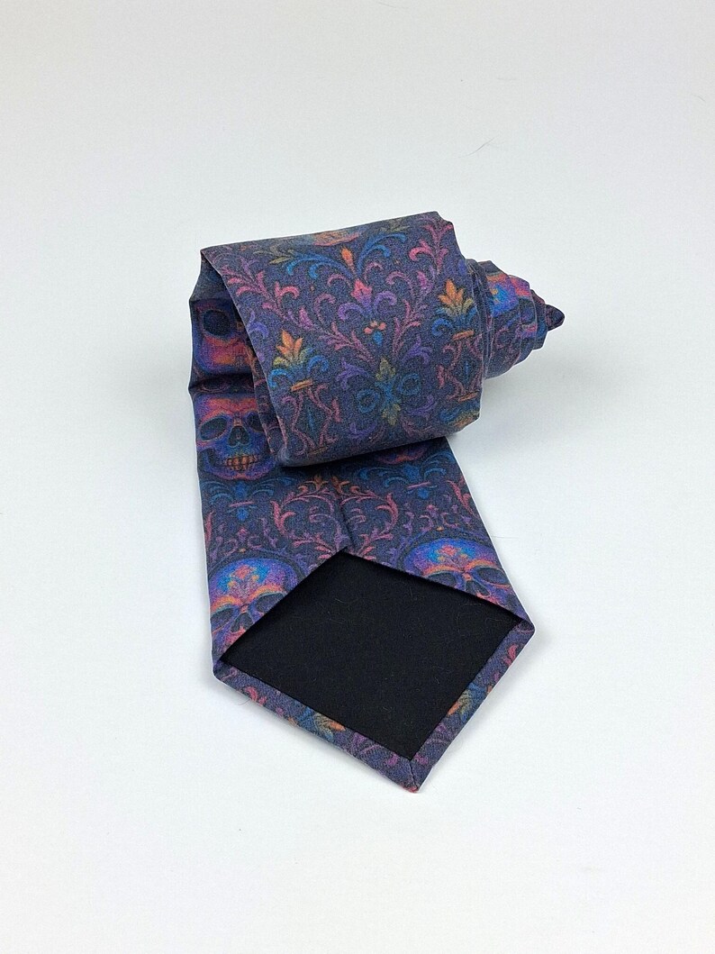 May include: A rolled-up necktie with a dark blue background and a repeating pattern of skulls and floral designs in shades of purple, orange, and blue. The tie has a black backing.