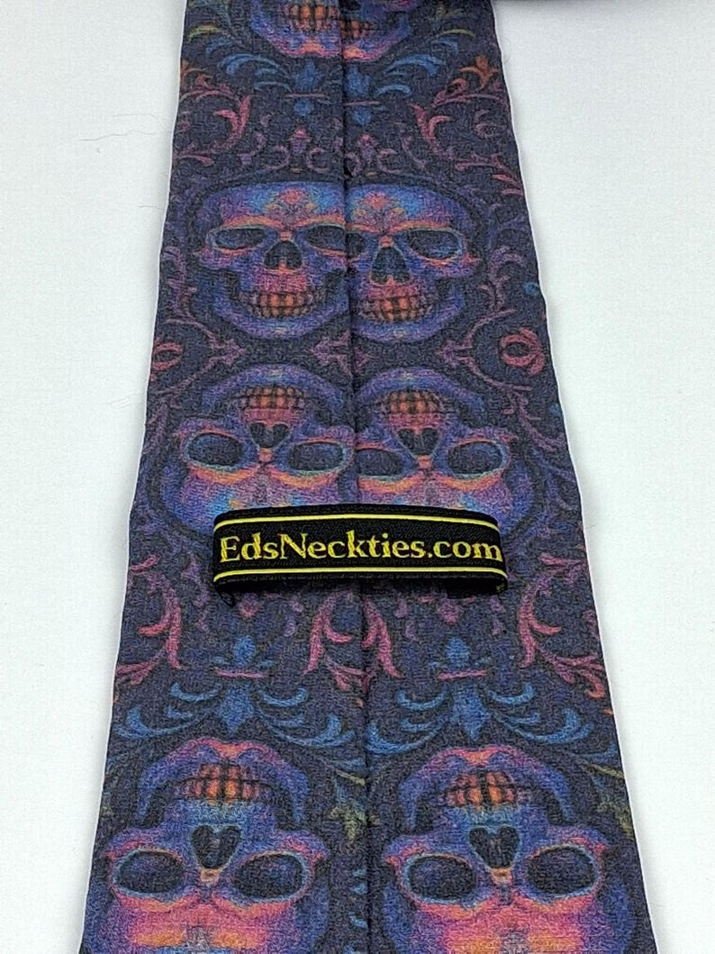 May include: A dark blue necktie with a repeating pattern of colourful skulls and floral designs. The tie has a black label with the text "EdsNeckties.com" in gold lettering. The tie is made of a smooth fabric.