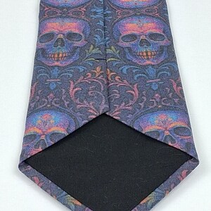 May include: A necktie featuring a repeating skull design in purple, blue, and orange hues. The tie has a dark navy blue backdrop with floral details. The reverse side of the tie is plain black.