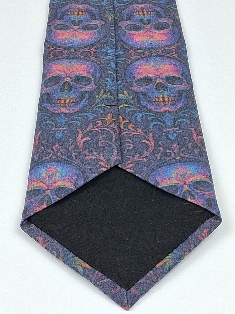 May include: A necktie featuring a repeating skull design in purple, blue, and orange hues. The tie has a dark navy blue backdrop with floral details. The reverse side of the tie is plain black.