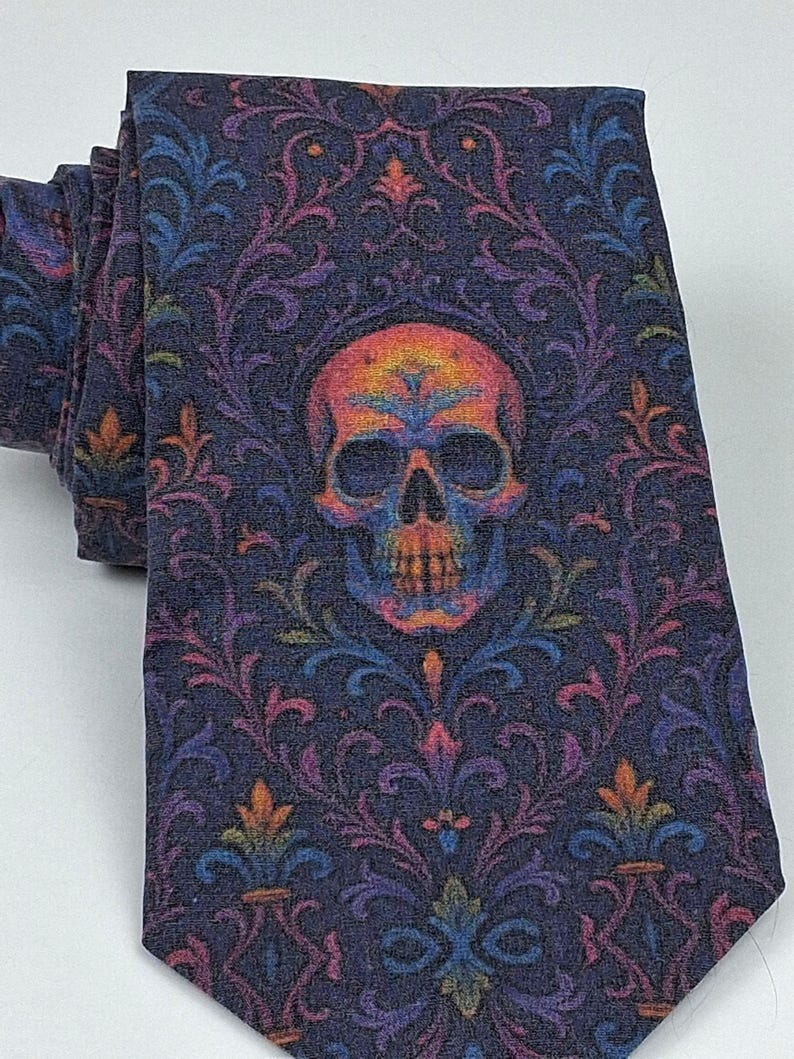 May include: A rolled-up tie featuring a dark blue backdrop and a vibrant skull motif. The skull is centrally positioned, encircled by a floral design in hues of purple, orange, blue, and green. The tie is crafted from a soft material.
