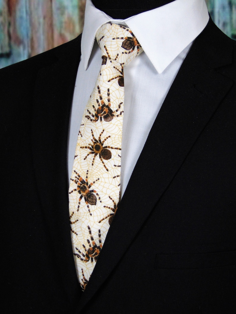 May include: A white tie with a pattern of brown spiders and webs. The tie is worn with a white dress shirt and a black suit jacket.