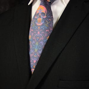 May include: A patterned necktie with a skull design and a colourful floral pattern. The tie is mainly grey with pink, orange, yellow, blue, and purple accents. The tie is worn with a white collared shirt and a black suit jacket.