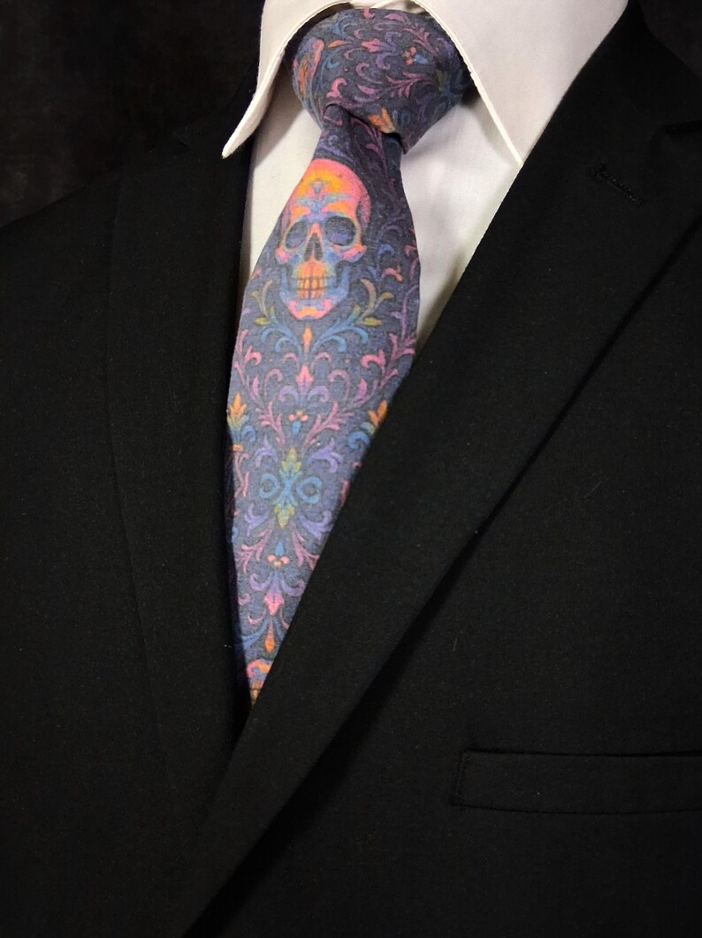 May include: A patterned necktie with a skull design and a colourful floral pattern. The tie is mainly grey with pink, orange, yellow, blue, and purple accents. The tie is worn with a white collared shirt and a black suit jacket.