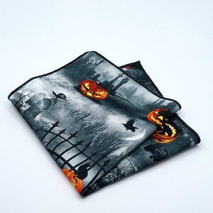 May include: A black and gray pocket square with a Halloween theme. It features a spooky graveyard scene with pumpkins, a crescent moon, and a raven.