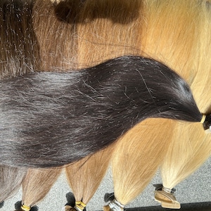 May include: A variety of human hair extensions in different shades of brown and blonde. The extensions are arranged in a row, with the darkest shade at the bottom and the lightest shade at the top. Each extension has a small, clear adhesive strip at the end.