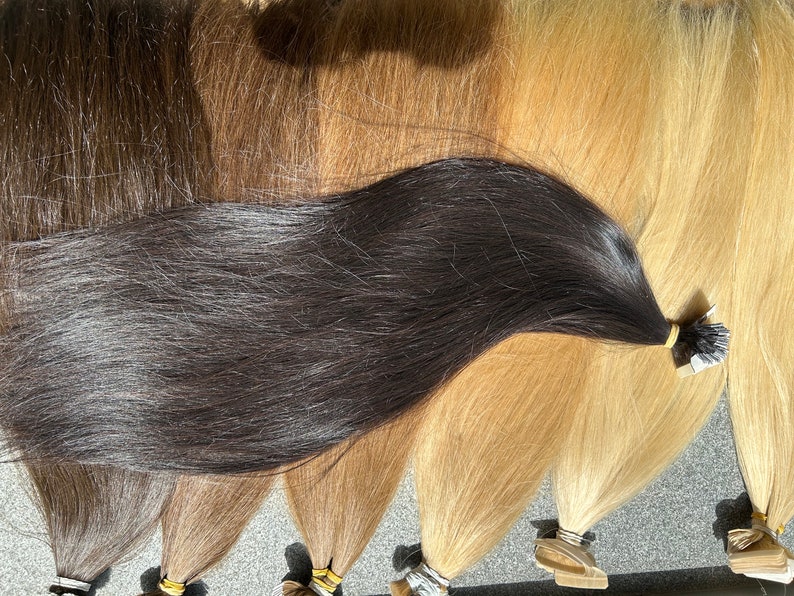 May include: A variety of human hair extensions in different shades of brown and blonde. The extensions are arranged in a row, with the darkest shade at the bottom and the lightest shade at the top. Each extension has a small, clear adhesive strip at the end.
