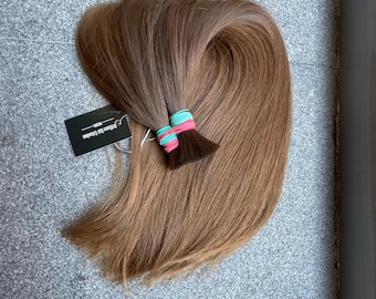 以下が含まれることがあります： ターコイズブルーとピンクのゴムバンドで束ねられたライトブラウンのヘアエクステンション。髪には小さな黒い長方形のタグが付いており、白い文字でBrilliance Hair ExtensionsとPONYという単語が記載されています。