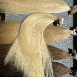 May include: Four wefts of human hair extensions in different shades of blonde and brown. The wefts are arranged in a row, with the lightest shade at the top and the darkest shade at the bottom.