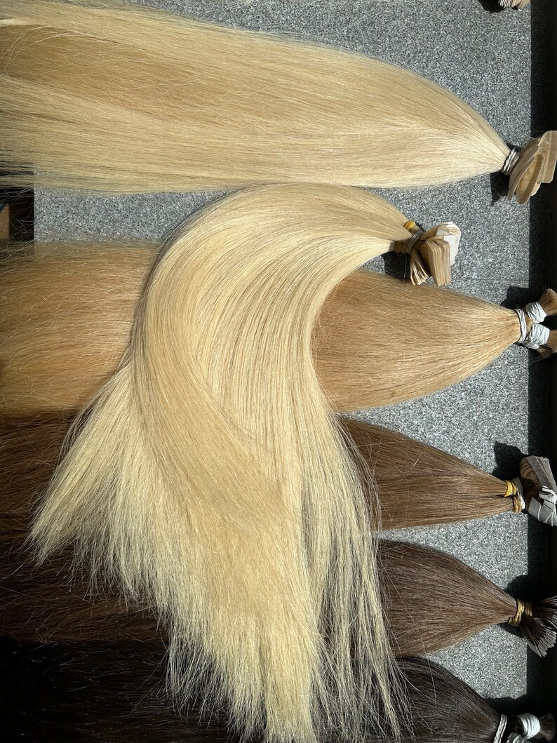 May include: Four wefts of human hair extensions in different shades of blonde and brown. The wefts are arranged in a row, with the lightest shade at the top and the darkest shade at the bottom.