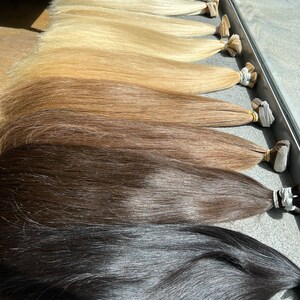 May include: A set of ten wefts of human hair extensions in various shades of blonde, brown, and black. The wefts are arranged in order of lightness, with the lightest shade at the top and the darkest shade at the bottom.
