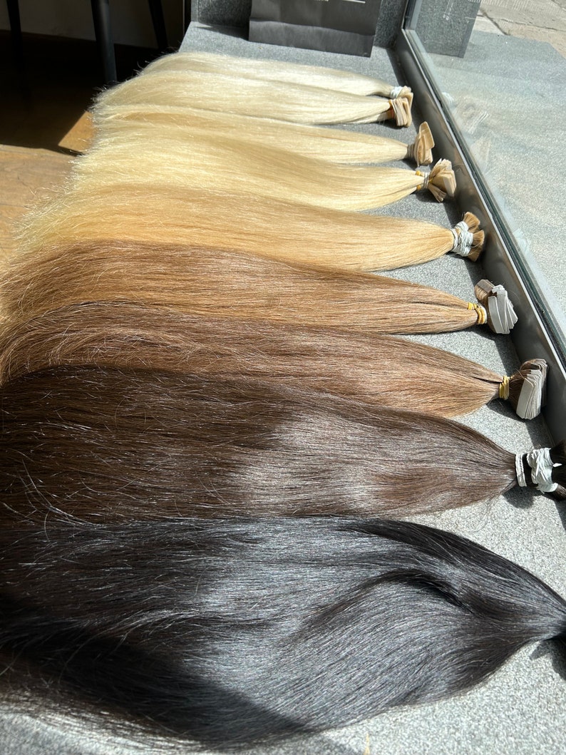 May include: A set of ten wefts of human hair extensions in various shades of blonde, brown, and black. The wefts are arranged in order of lightness, with the lightest shade at the top and the darkest shade at the bottom.