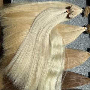 May include: Three bundles of human hair extensions in different shades of blonde. The top bundle is the lightest blonde, the middle bundle is a medium blonde, and the bottom bundle is a dark blonde. The hair extensions are all straight and have a natural shine.