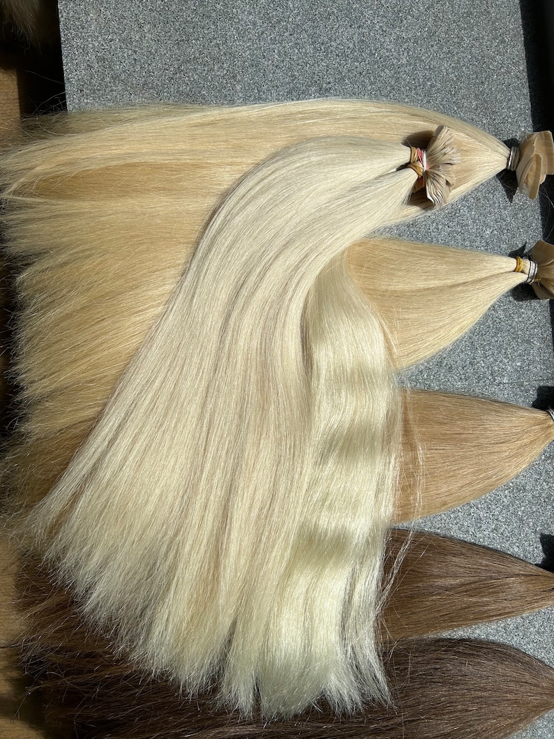 May include: Three bundles of human hair extensions in different shades of blonde. The top bundle is the lightest blonde, the middle bundle is a medium blonde, and the bottom bundle is a dark blonde. The hair extensions are all straight and have a natural shine.