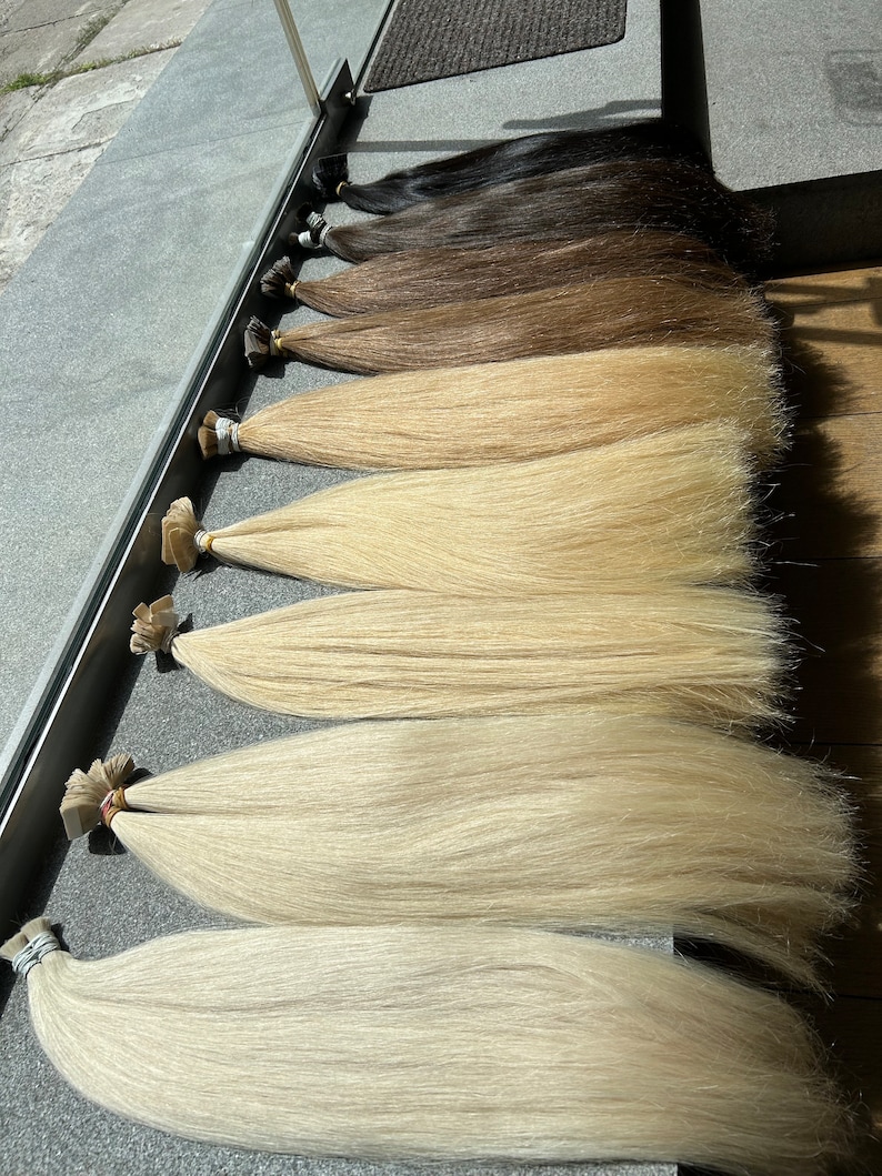 May include: A variety of human hair extensions in different shades of blonde and brown, arranged in a row from darkest to lightest. The extensions are long and straight, with a smooth texture.