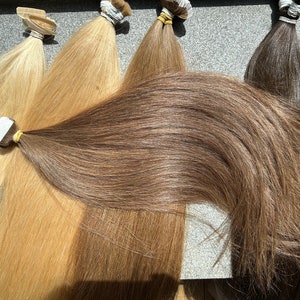 May include: Five wefts of human hair extensions in various shades of blonde and brown. The wefts are arranged horizontally, with the darkest weft at the bottom and the lightest weft at the top.