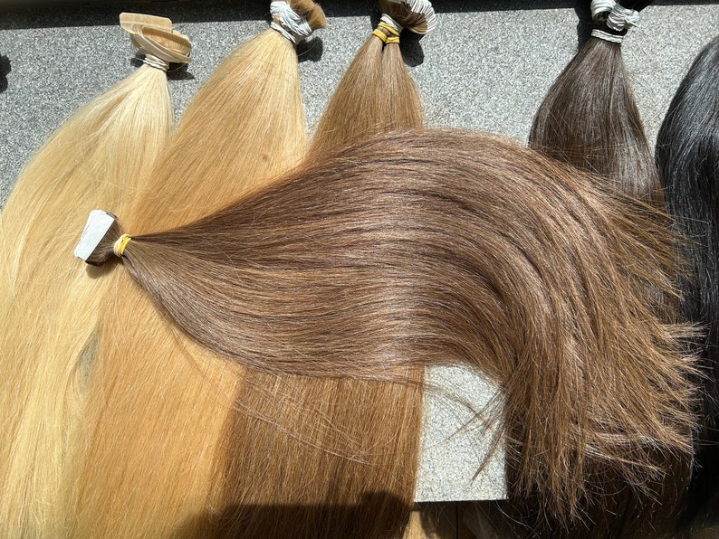 May include: Five wefts of human hair extensions in various shades of blonde and brown. The wefts are arranged horizontally, with the darkest weft at the bottom and the lightest weft at the top.