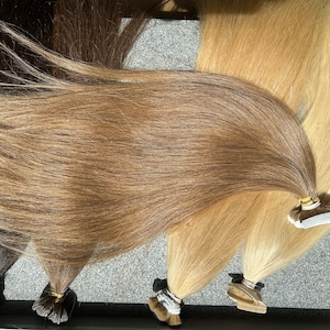 May include: A set of four wefts of human hair extensions in various shades of brown and blonde. The wefts are arranged in a fan shape and have tape attachments.
