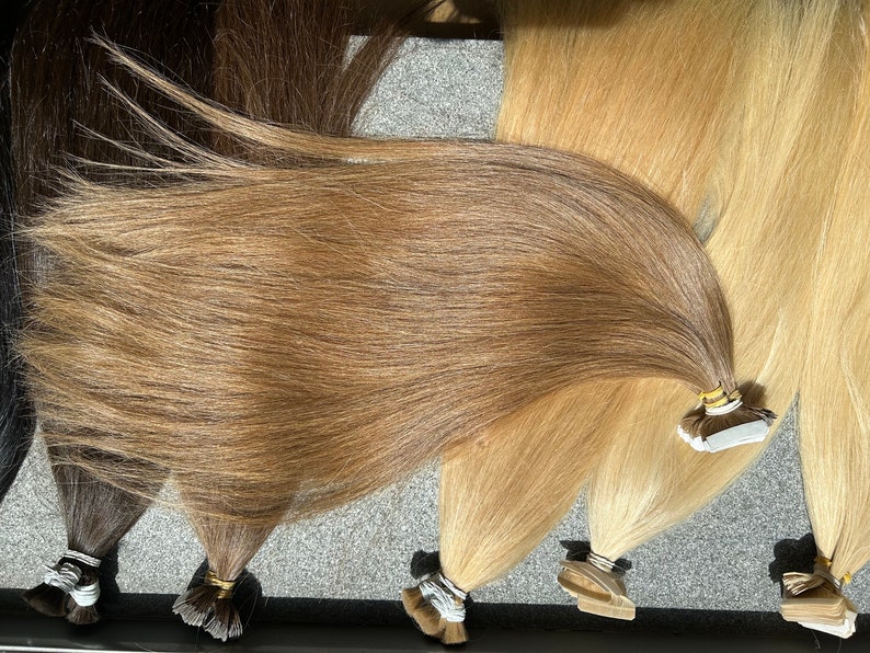 May include: A set of four wefts of human hair extensions in various shades of brown and blonde. The wefts are arranged in a fan shape and have tape attachments.
