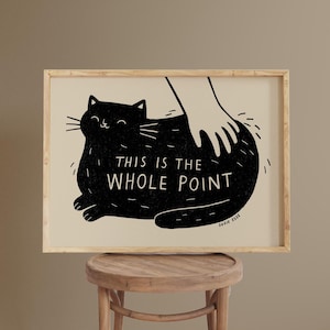 Peut inclure: Une affiche encadrée représentant un chat noir caressé par une main. Le texte "THIS IS THE WHOLE POINT" est affiché en dessous. L'œuvre est encadrée dans un cadre en bois et posée sur un tabouret.