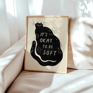 Peut inclure: Une affiche encadrée avec une illustration de chat noir. Le corps du chat forme une forme fluide, avec les mots "IT'S OKAY TO BE SOFT" au centre. L'œuvre est sur fond crème, encadrée dans un fin cadre doré.