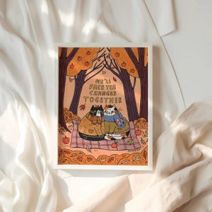 Peut inclure: Une impression d'art encadrée représentant deux chats blottis sur une couverture de pique-nique sous des arbres d'automne. L'œuvre d'art a une palette de couleurs chaudes avec le texte "We'll Face The Changes Together".