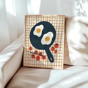 May include: A framed art print featuring a dark blue frying pan with two smiling fried eggs inside, surrounded by red cherry tomatoes on a vine, set against a background of a tan and white grid pattern. The print is enclosed in a thin gold frame.