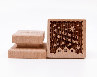 May include: A wooden stamp with a Christmas design. The stamp features a snowflake pattern, the words "Frohliche Weihnachten", and three small houses.