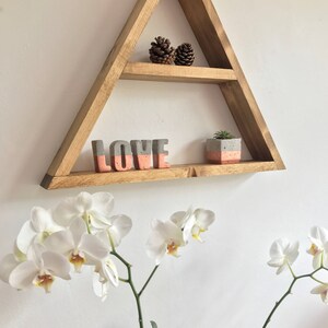 Puede incluir: Una estanter&iacute;a triangular de madera con dos niveles, que contiene objetos decorativos. La estanter&iacute;a muestra dos pi&ntilde;as, la palabra "LOVE" en letras grises y coral, y una peque&ntilde;a suculenta en maceta. Orqu&iacute;deas blancas con centros amarillos en primer plano.