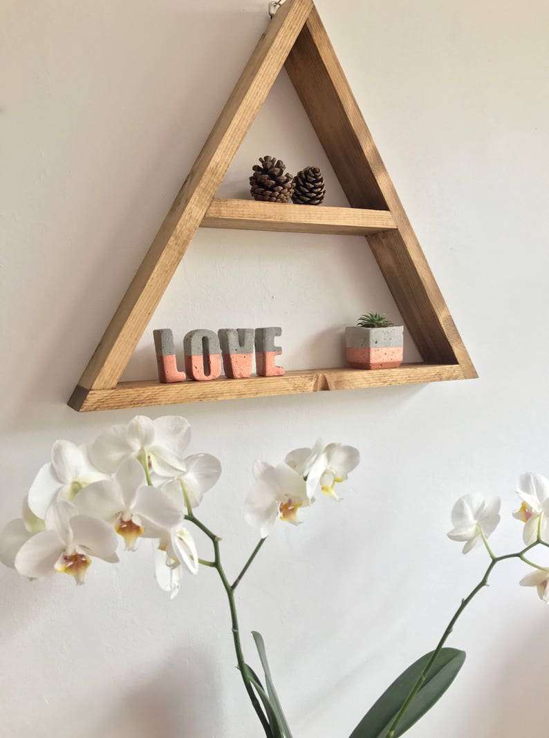 Puede incluir: Una estanter&iacute;a triangular de madera con dos niveles, que contiene objetos decorativos. La estanter&iacute;a muestra dos pi&ntilde;as, la palabra "LOVE" en letras grises y coral, y una peque&ntilde;a suculenta en maceta. Orqu&iacute;deas blancas con centros amarillos en primer plano.