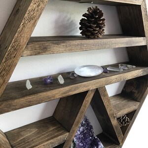 May include: A hexagonal wooden shelf with multiple shelves and compartments. The shelf is stained a dark brown color and has a rustic look. The shelf is decorated with crystals, pine cones, and a large amethyst geode.