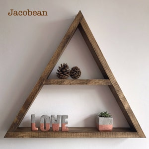 Puede incluir: Estanter&iacute;a triangular de madera con dos niveles. Contiene dos pi&ntilde;as, la palabra "LOVE" en letras grises y coral, y una peque&ntilde;a suculenta en maceta. La estanter&iacute;a es de color marr&oacute;n c&aacute;lido y est&aacute; montada en una pared blanca.