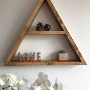 Puede incluir: Estanter&iacute;a triangular de madera con dos niveles, que contiene dos pi&ntilde;as, letras de hormig&oacute;n que deletrean "LOVE" con un acento coral y una peque&ntilde;a suculenta en maceta. La palabra "Georgian" es visible.