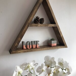 Puede incluir: Una estanter&iacute;a triangular de madera marr&oacute;n con dos niveles. La balda superior contiene dos pi&ntilde;as. La balda inferior muestra una escultura de hormig&oacute;n con la palabra "LOVE" en gris y coral, y una peque&ntilde;a planta en maceta.