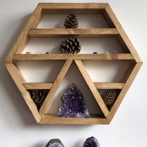 May include: A hexagonal wooden shelf with five shelves, featuring a large amethyst crystal and pine cones. The shelf is designed to display crystals and other small objects.