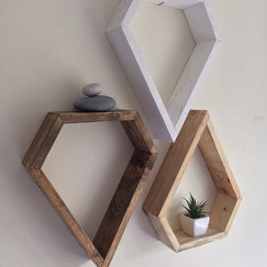 May include: Three diamond-shaped shelves, one white and two stained wood, mounted on a wall. The top shelf is empty, the middle shelf has two stacked gray stones, and the bottom shelf has a small potted plant.