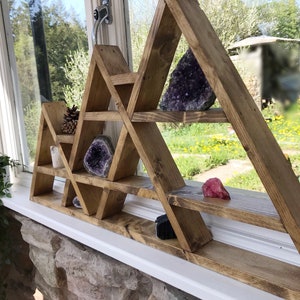 Puede incluir: Estantería triangular de madera con múltiples estantes, diseñada para exhibir cristales y otros objetos pequeños. La estantería está teñida de un color marrón oscuro y tiene un aspecto rústico. Está montada en una pared blanca y presenta una variedad de cristales, incluyendo amatista, cuarzo rosa y obsidiana negra.