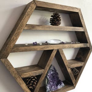 May include: A hexagonal wooden shelf with multiple compartments, featuring a large amethyst crystal, pine cones, and small clear and purple crystals. The shelf is stained a dark brown color and has a rustic, farmhouse style.