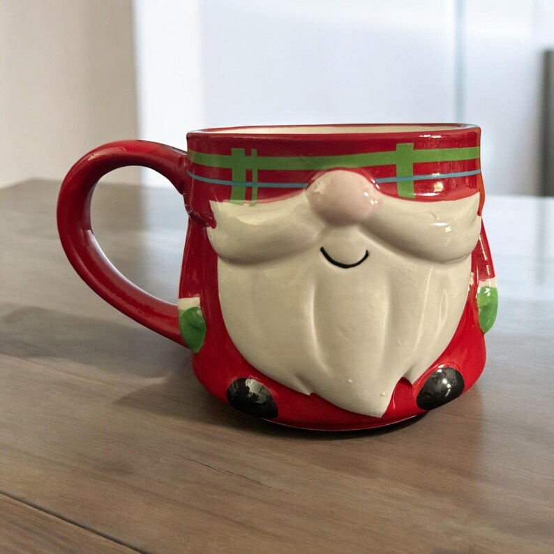Peut inclure: Mug en c&eacute;ramique rouge en forme de P&egrave;re No&euml;l avec une barbe blanche et un chapeau vert. Le mug a un motif &agrave; carreaux autour du haut.