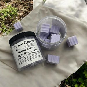 May include: A glass jar filled with lavender-colored sugar scrub cubes. The jar has a black lid and a label that reads "Ivy Creek Wisteria & Lilacs Sugar Scrub Cubes Exfoliating Gentle Single Use".