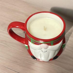 Peut inclure: Une tasse en c&eacute;ramique rouge et blanche en forme de P&egrave;re No&euml;l tenant une bougie. La bougie est blanche et a une m&egrave;che.