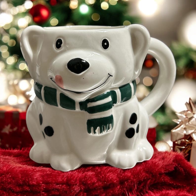 May include: White ceramic mug shaped like a dog wearing a green and white scarf. The mug has black spots and a pink tongue sticking out.