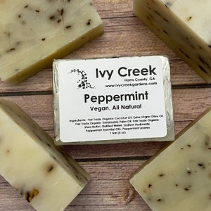 May include: A close-up of several bars of handmade peppermint soap. The soap is light beige with dark specks and a label that reads "Peppermint, Vegan, All Natural" from Ivy Creek Gardens. The label also lists the ingredients.