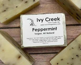 May include: A close-up of several bars of handmade peppermint soap. The soap is light beige with dark specks and a label that reads "Peppermint, Vegan, All Natural" from Ivy Creek Gardens. The label also lists the ingredients.